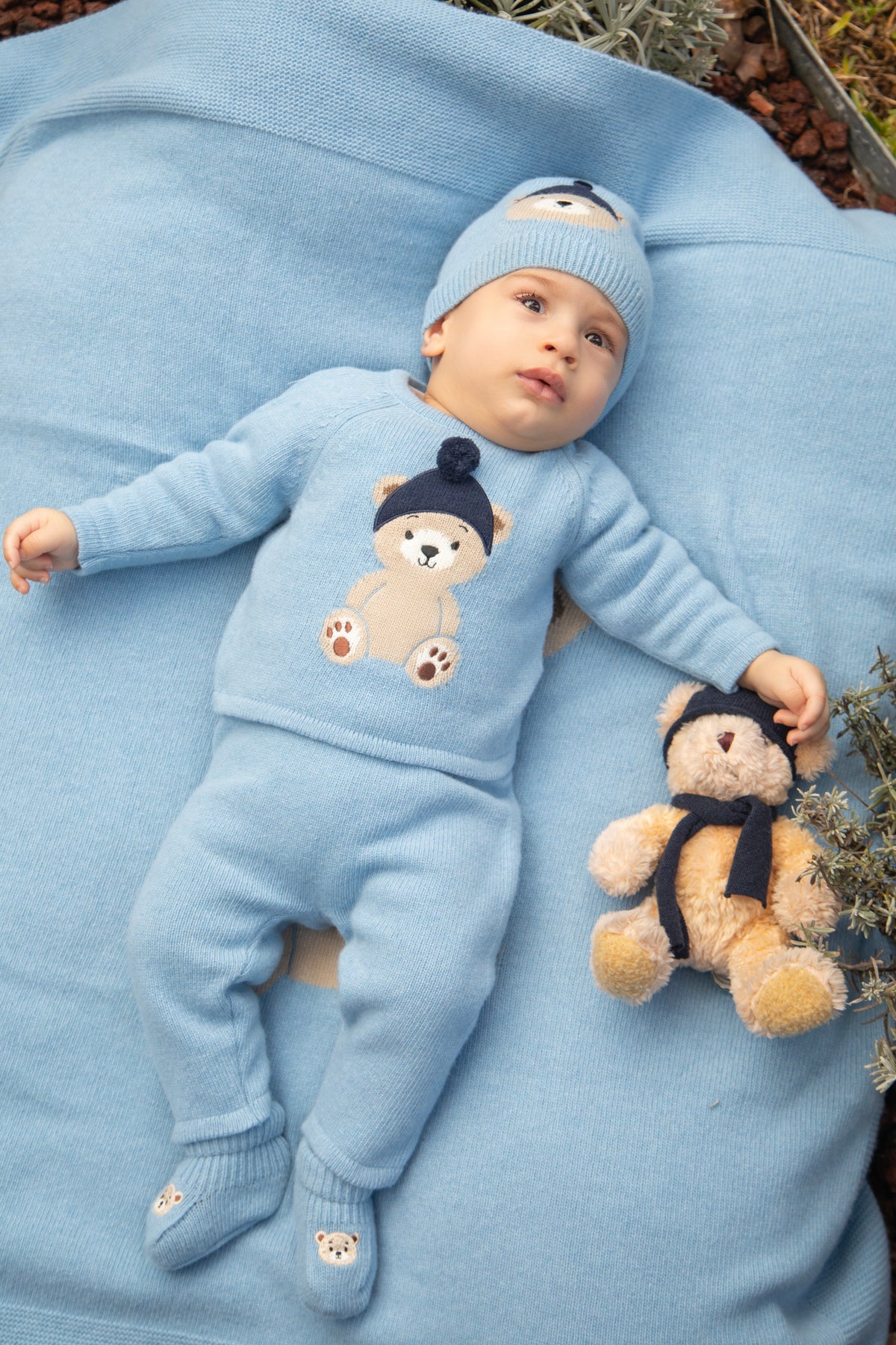 Baby in lichtblauw gebreid babypakje met teddybeer motief en bijpassend mutsje – sfeerbeeld