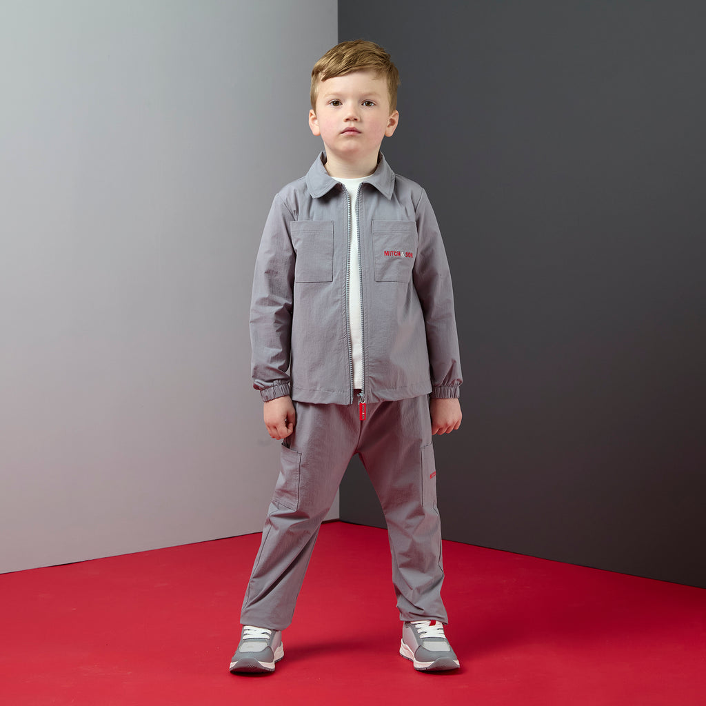 Jongen in grijs 2-delig Thiago pak van Mitch & Son in parachutestof met rits en rood logo, gedragen met sneakers
